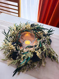 A dried floral wreath centerpiece with green leaves, small cream flowers, and a lit glass tealight candle, displayed on a beige table runner with a window and orange curtain in the background.
