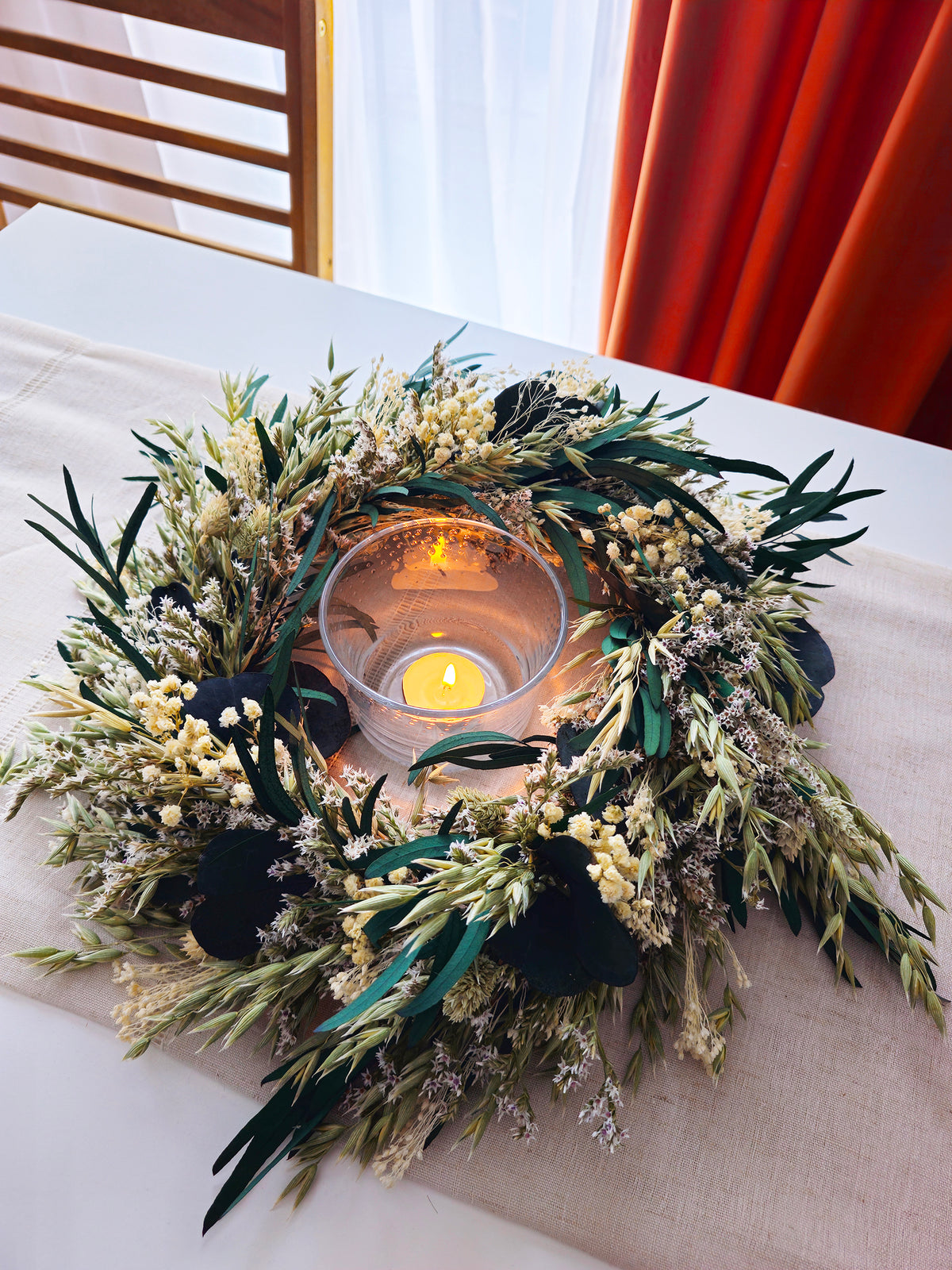 A dried floral wreath centerpiece with green leaves, small cream flowers, and a lit glass tealight candle, displayed on a beige table runner with a window and orange curtain in the background.