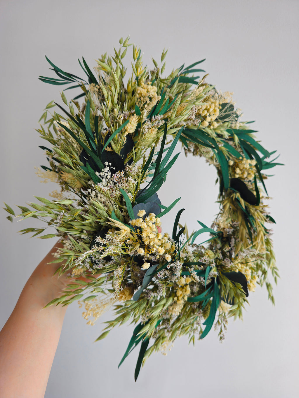 A dried flower wreath with green and ivory foliage, small cream flowers, and dark green accents, held by a person's hand against a plain light gray background. It includes eucalyptus, baby's breath and other grasses.