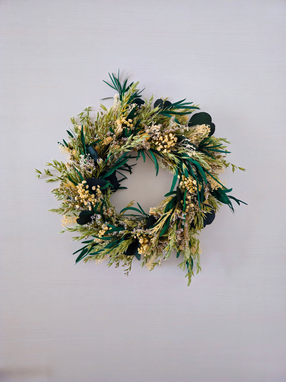 Green advent wreath displayed on a white wall. The wreath features eucalyptus, dried grasses and white gypsophila.