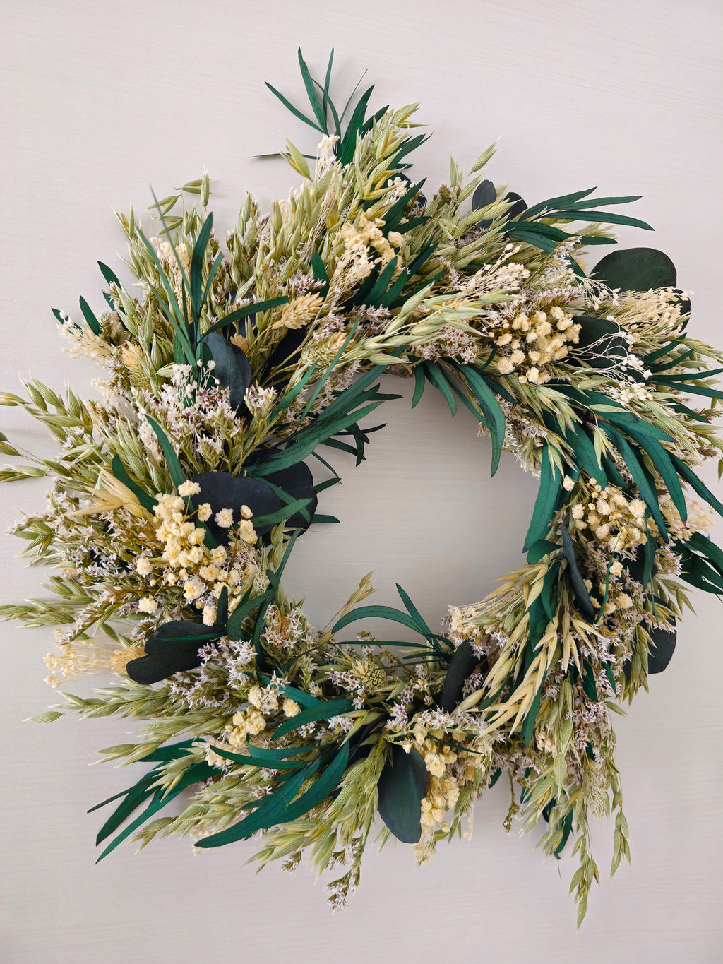 Green advent wreath displayed on a white wall. The wreath features eucalyptus, dried grasses and white gypsophila.