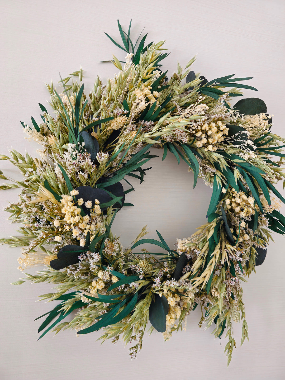 Green advent wreath displayed on a white wall. The wreath features eucalyptus, dried grasses and white gypsophila.