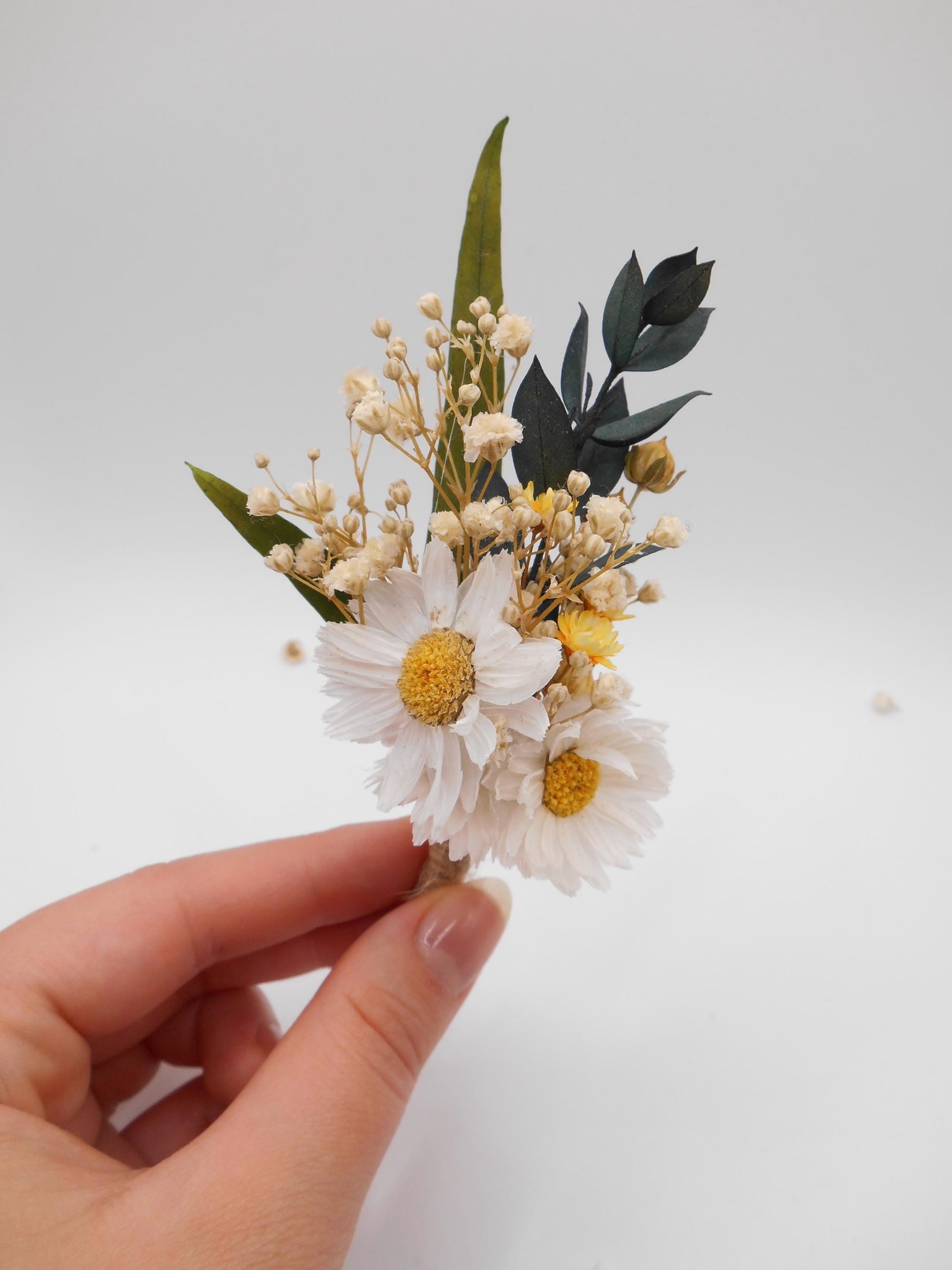 Hand holding a boutonniere with white daisies and yellow flowers, green leaves, and eucalyptus on a white background
