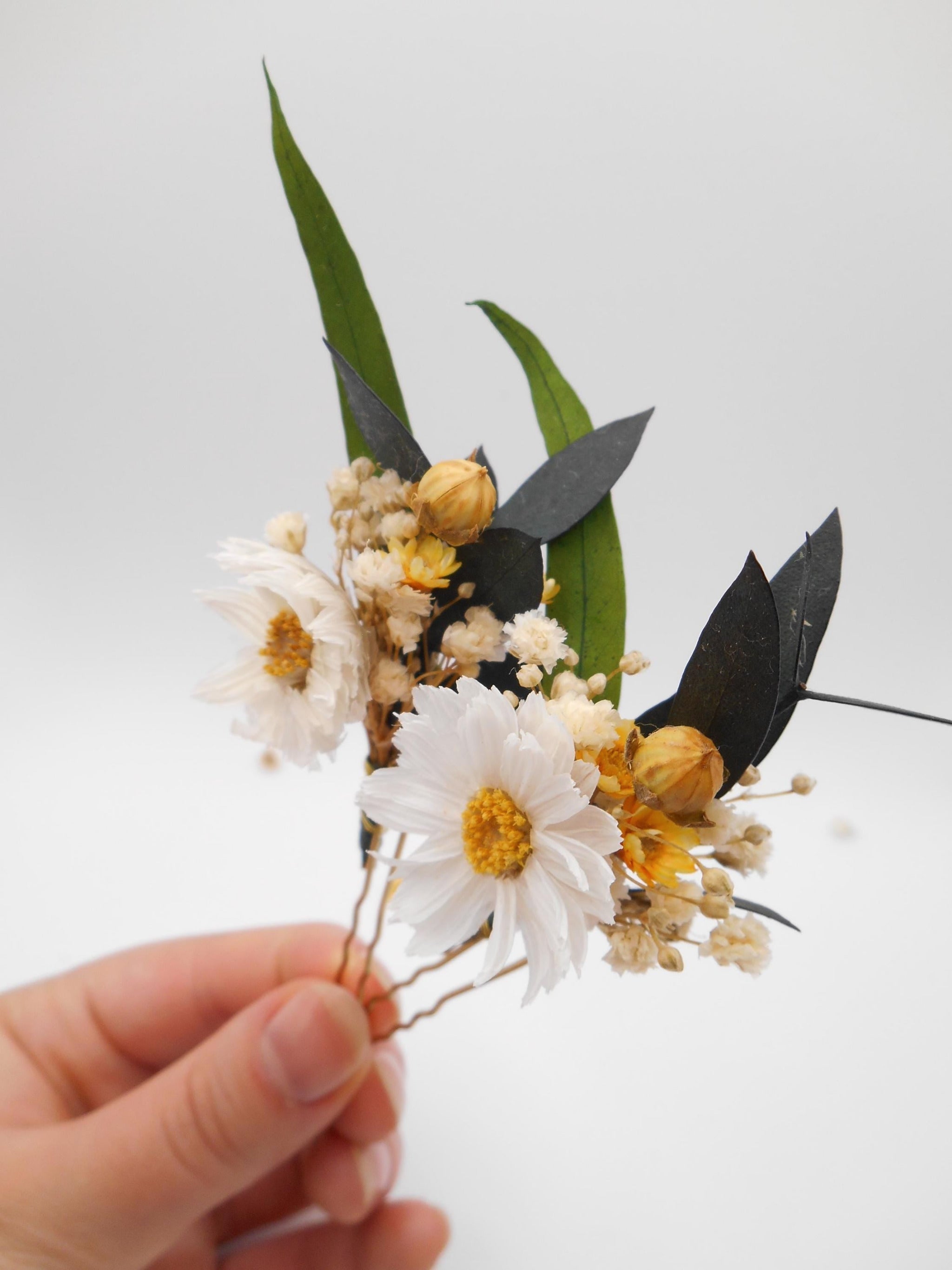 Hand holding two floral hairpins with white daisies and yellow  flowers, green leaves, and gold accents on a white background